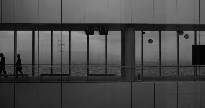 a grayscale photo of an office corridor with glass walls, with two men walking in it