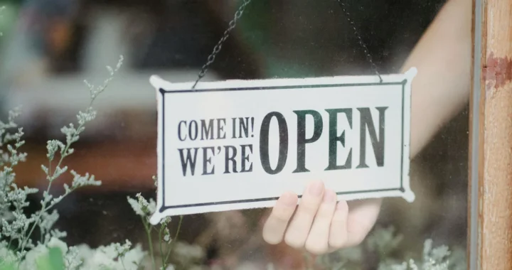 a hand holding on an open sign attached on a glass door