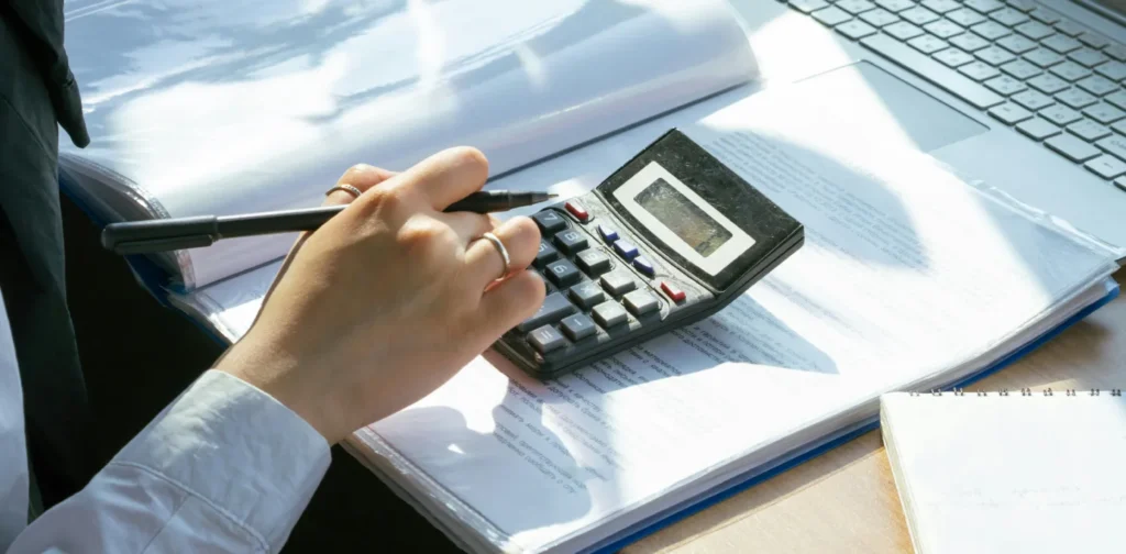 hands of a person trying to an esg report using a calculator on top of documents in front of a laptop