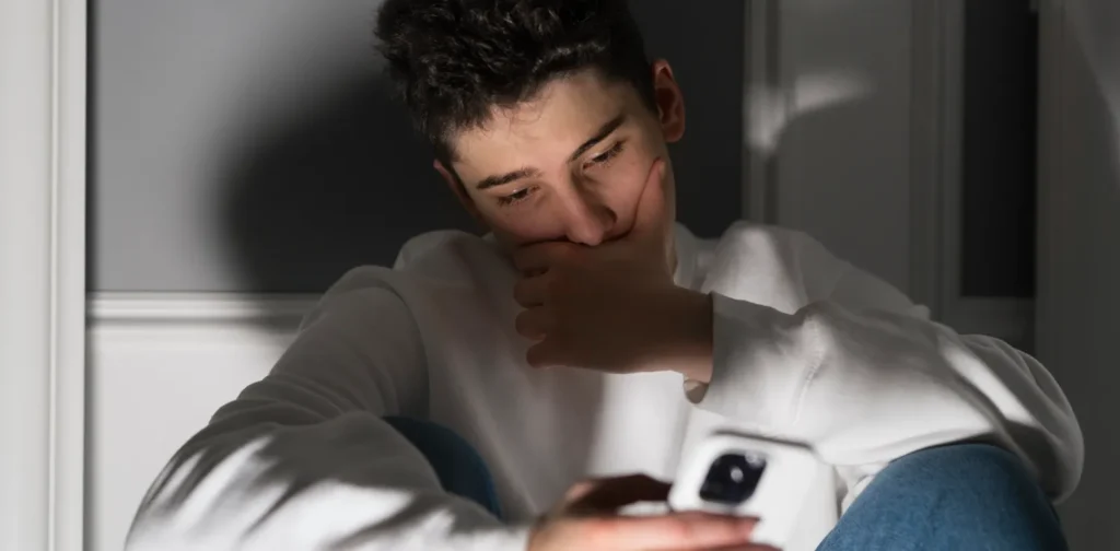 A young man sits alone in a low light room, looking distressed while holding his phone.