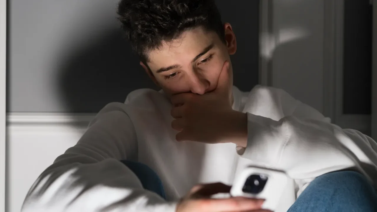 A young man sits alone in a low light room, looking distressed while holding his phone.