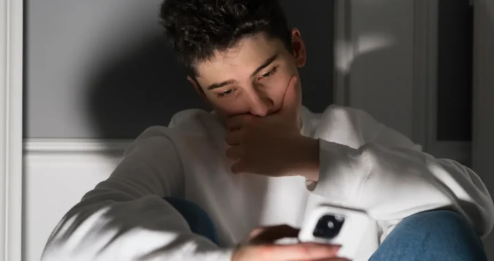 A young man sits alone in a low light room, looking distressed while holding his phone.
