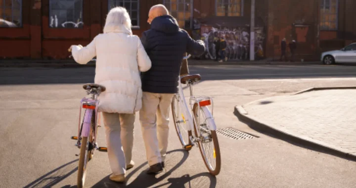 An elderly couple walks side by side while holding their bicycles on a quiet street.