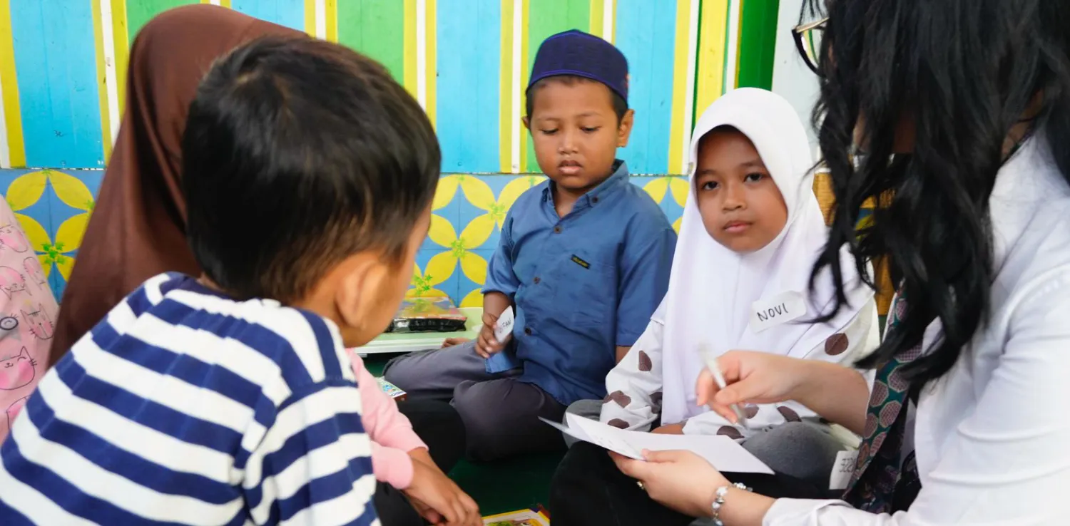 a woman volunteer and four children reading and writing together in a circle at the Inisiasi lantara literacy event