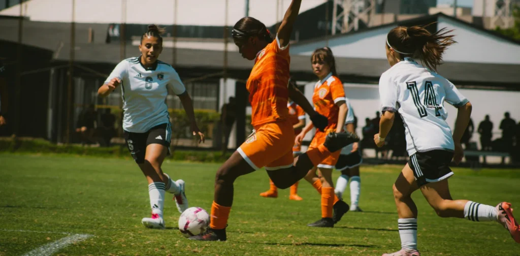 A female soccer player in orange kicks the ball while competing against opponents in white jerseys during a match.