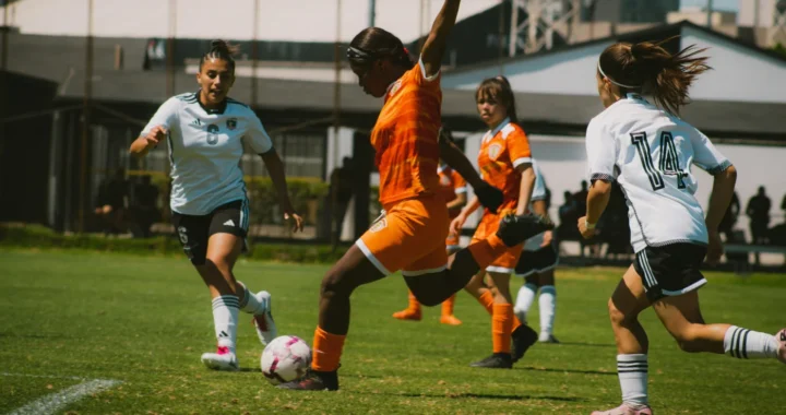 A female soccer player in orange kicks the ball while competing against opponents in white jerseys during a match.