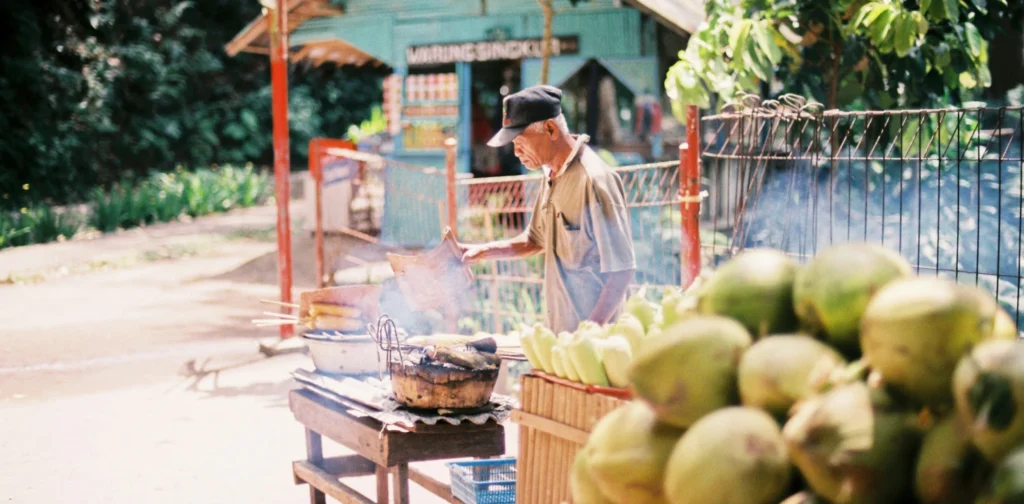 A man selling and fanning grilled corns beside a table full of young coconuts.