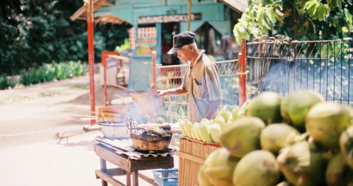 A man selling and fanning grilled corns beside a table full of young coconuts.