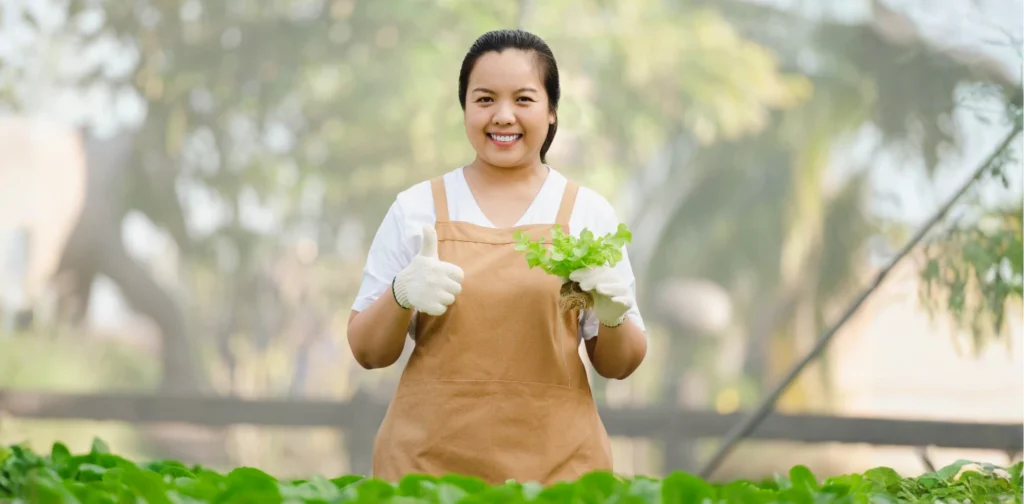 A female farmer holding fresh vegetables in a greenhouse, smiling and giving a thumbs-up.