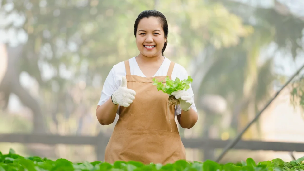 A female farmer holding fresh vegetables in a greenhouse, smiling and giving a thumbs-up.