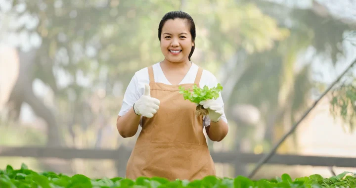 A female farmer holding fresh vegetables in a greenhouse, smiling and giving a thumbs-up.