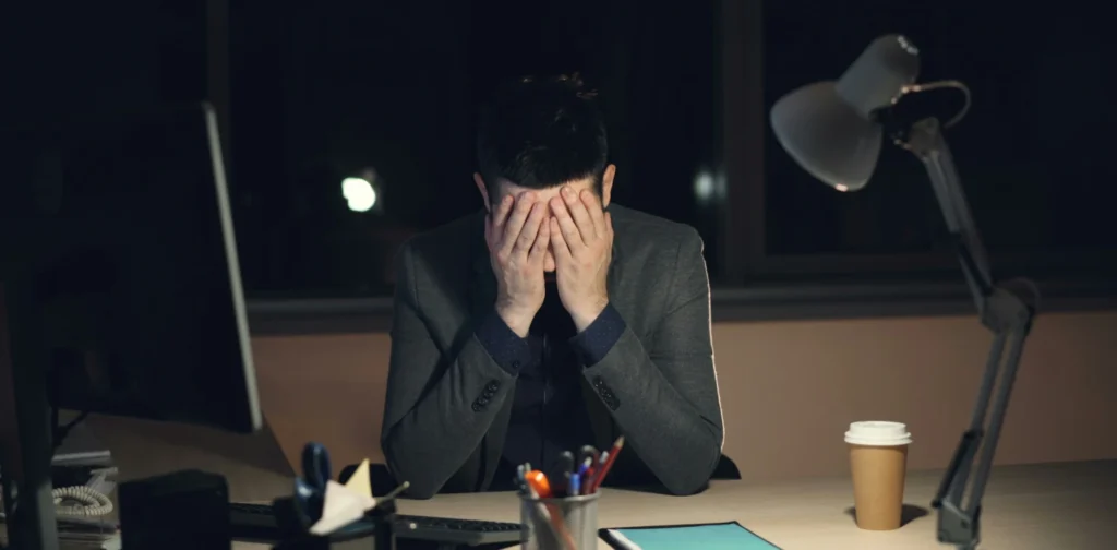 Tired-looking young man sits in front of a computer with his head in between his hands.