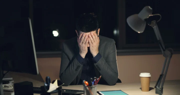 Tired-looking young man sits in front of a computer with his head in between his hands.