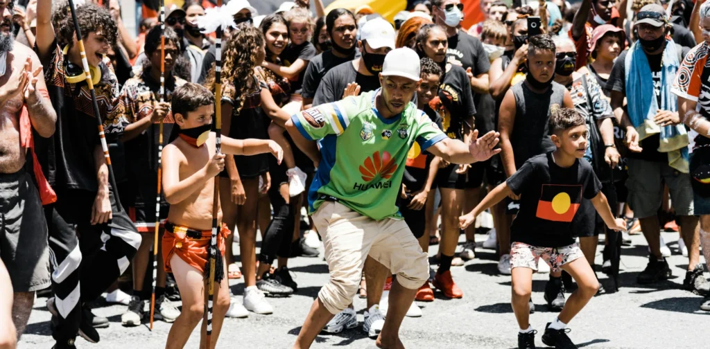 some Aboriginal peoples–kids and adults–wearing their traditional attributes in the middle of a rally in the street, with Aboriginal flags visible in the background
