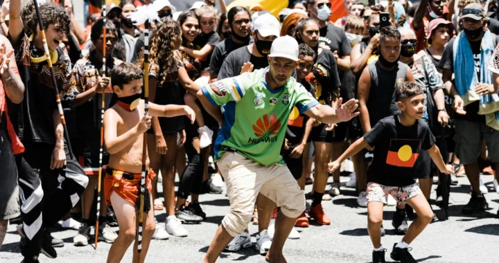 some Aboriginal peoples–kids and adults–wearing their traditional attributes in the middle of a rally in the street, with Aboriginal flags visible in the background