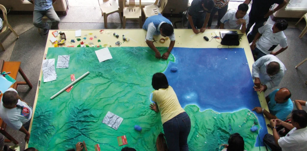 an aerial photo of people gathering around a huge table with geography model on it