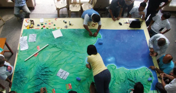 an aerial photo of people gathering around a huge table with geography model on it