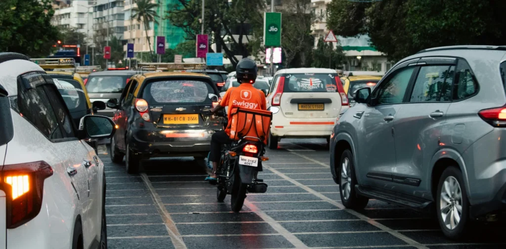 A delivery rider in a bright orange jacket navigates through dense Indian traffic on his motorbike.