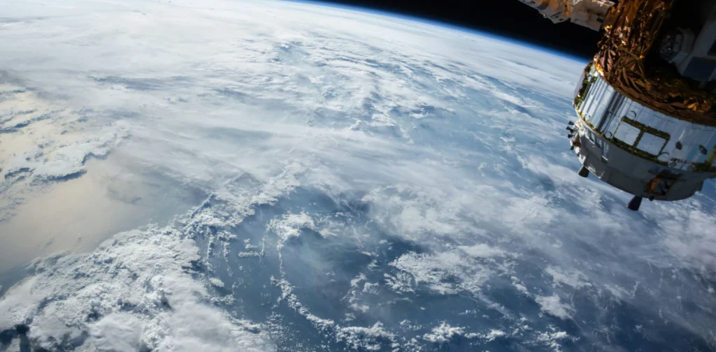 View of earth from orbit, showing the planet’s horizon, blue ocean, and swirling white cloud, with part of a satellite visible at the right edge.