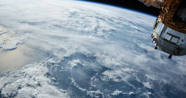 View of earth from orbit, showing the planet’s horizon, blue ocean, and swirling white cloud, with part of a satellite visible at the right edge.