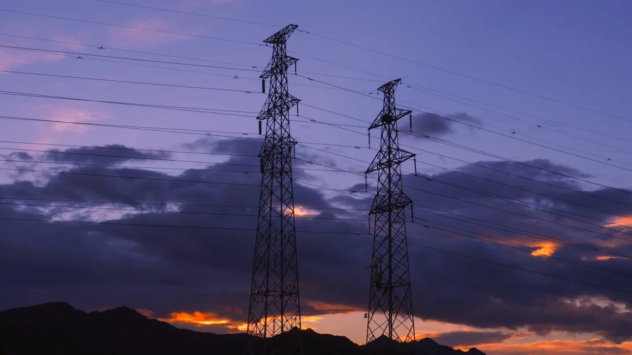 Two tall electricity transmission towers silhouetted against a purple and orange sunset sky, with power lines stretching across dark clouds and distant mountains.