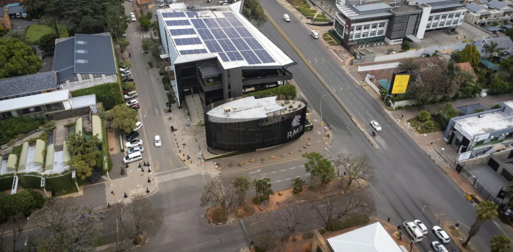 Aerial view of an urban intersection in south africa with modern buildings, one with rooftop solar panels, and light traffic on surrounding streets.