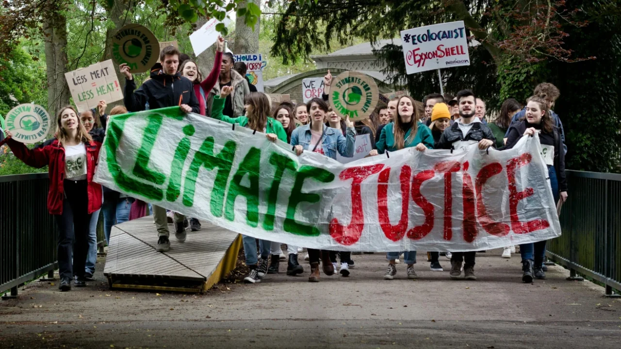 A group of young climate activists marching across a bridge holding a large, “Climate Justice” banner and protest signs during a climate strike.