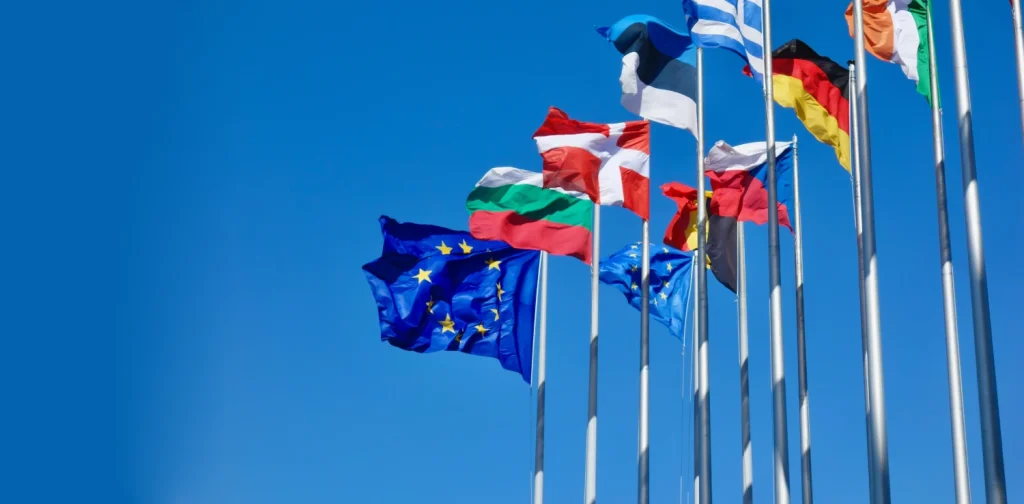 Multiple European flags, including the European Union flag, flying on tall flagpoles against a clear blue sky.