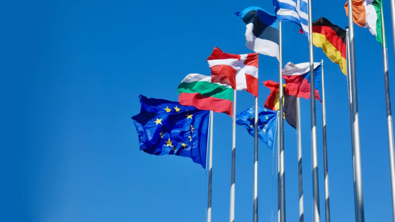 Multiple European flags, including the European Union flag, flying on tall flagpoles against a clear blue sky.