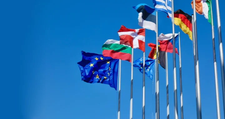Multiple European flags, including the European Union flag, flying on tall flagpoles against a clear blue sky.