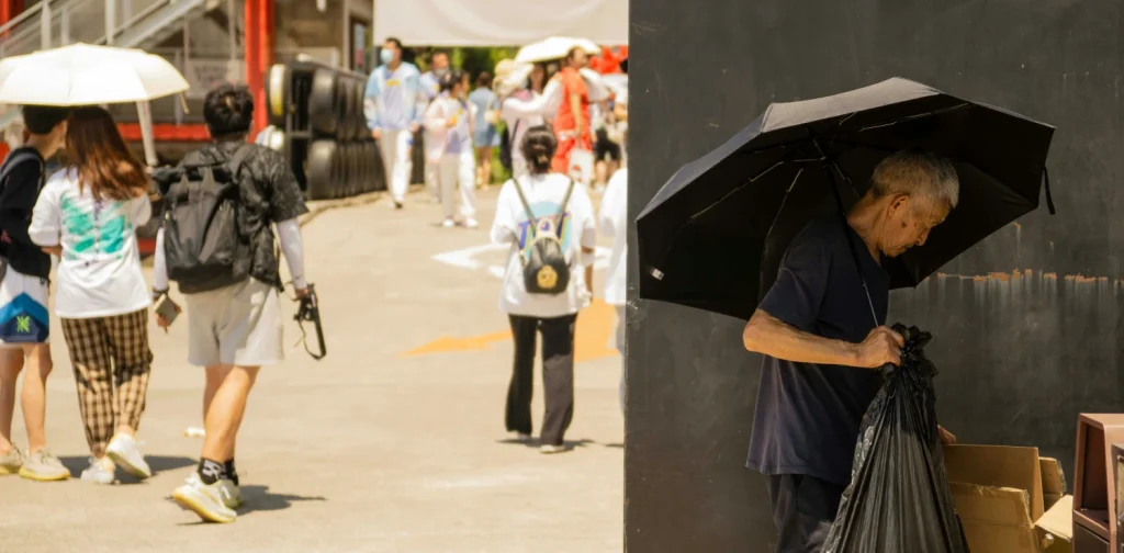 an old man standing on the side of a busy, sunny street. he is sorting through cardboard while holding a black umbrella.