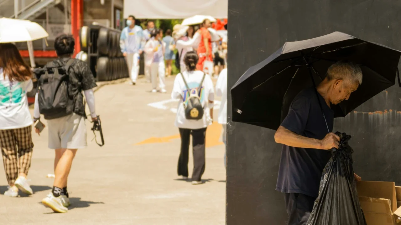 an old man standing on the side of a busy, sunny street. he is sorting through cardboard while holding a black umbrella.