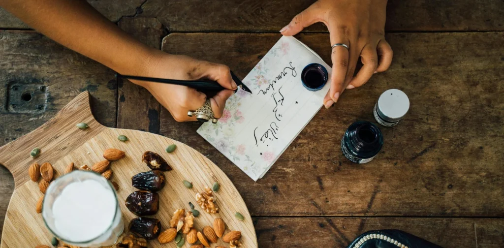 person writing on a paper on a table set with a tray of dates and nuts and a glass of milk