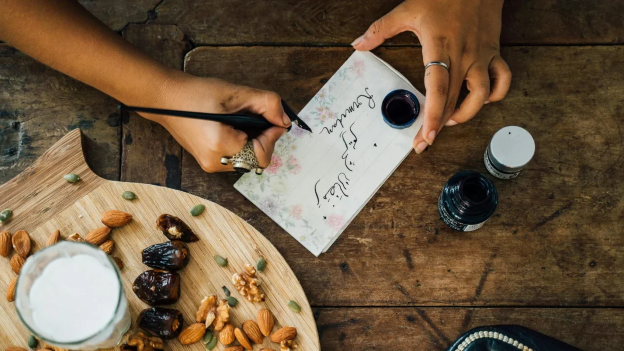 person writing on a paper on a table set with a tray of dates and nuts and a glass of milk