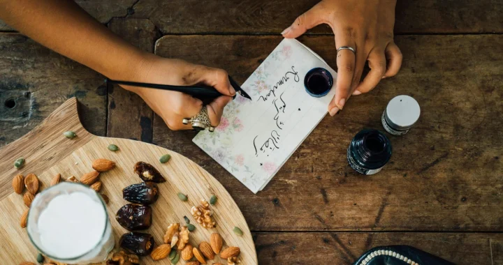 person writing on a paper on a table set with a tray of dates and nuts and a glass of milk