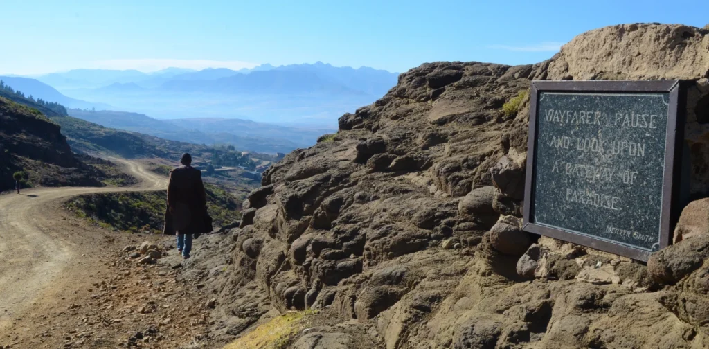 A person walks along a dirt mountain road overlooking the vast highland landscape of Lesotho, with layered mountains in the distance and a stone plaque reading “Wayfarer, pause and look upon a pathway to paradise.”