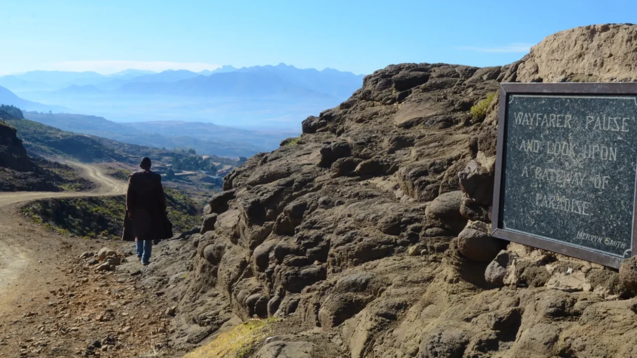 A person walks along a dirt mountain road overlooking the vast highland landscape of Lesotho, with layered mountains in the distance and a stone plaque reading “Wayfarer, pause and look upon a pathway to paradise.”