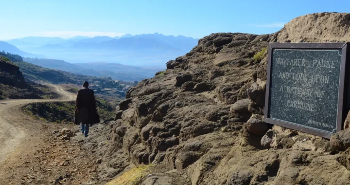 A person walks along a dirt mountain road overlooking the vast highland landscape of Lesotho, with layered mountains in the distance and a stone plaque reading “Wayfarer, pause and look upon a pathway to paradise.”