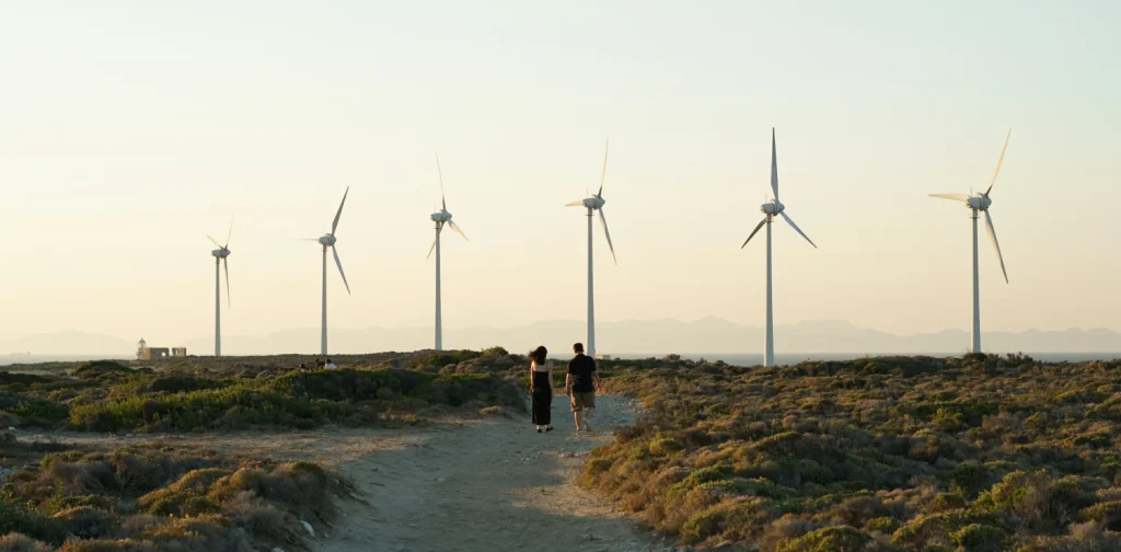a man and a woman walking down a path with dirt and greenery with wind turbines in the background