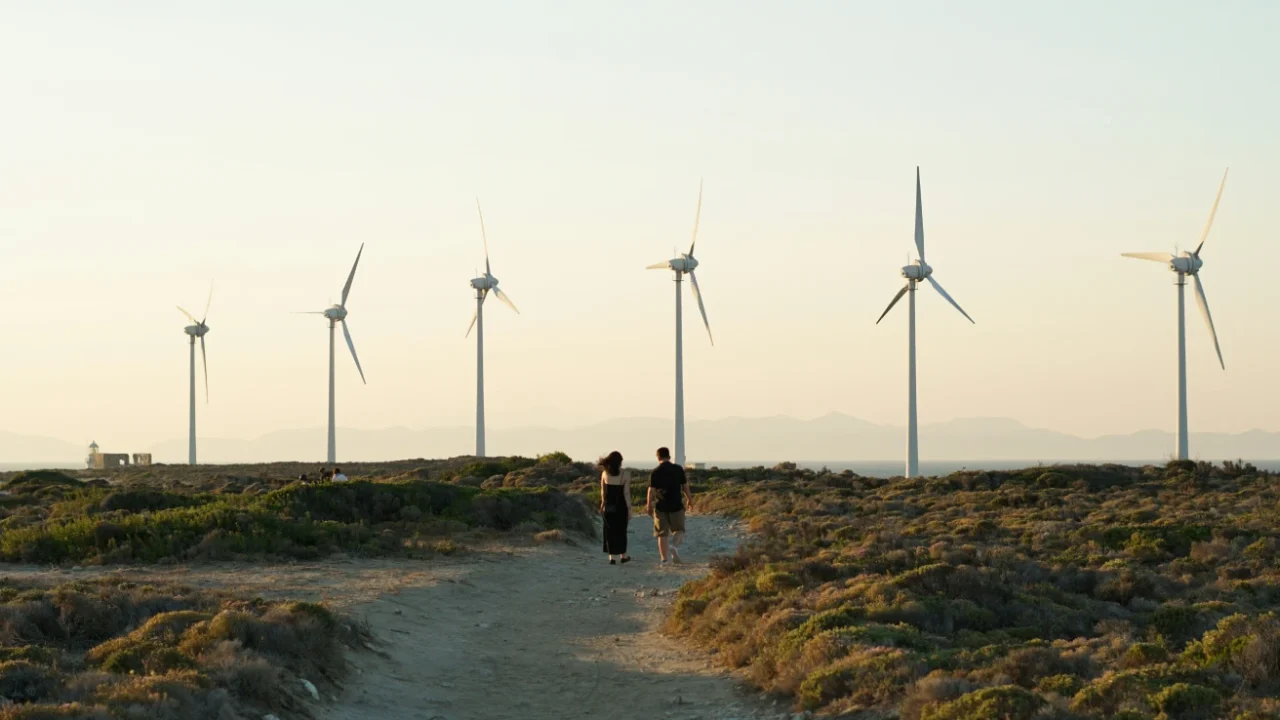 a man and a woman walking down a path with dirt and greenery with wind turbines in the background