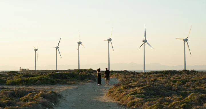 a man and a woman walking down a path with dirt and greenery with wind turbines in the background