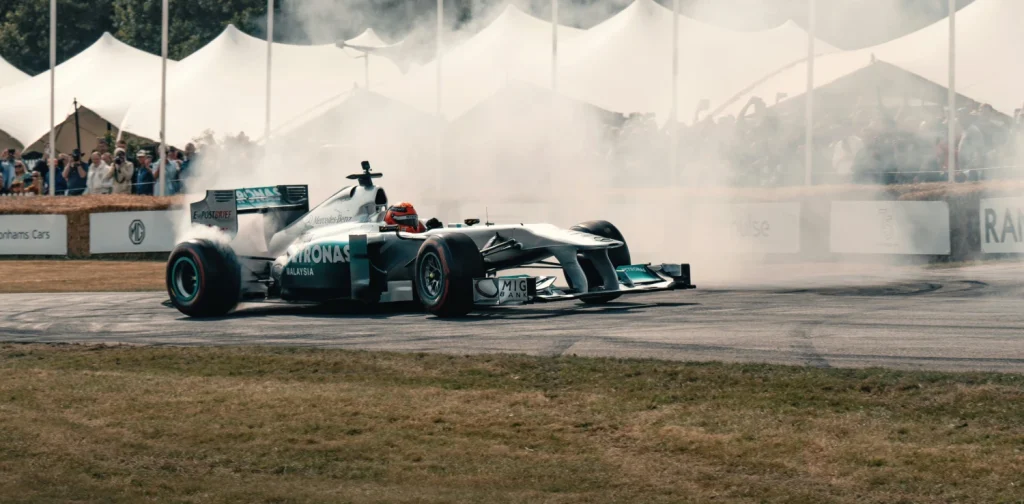 A silver Formula 1 race car performs a smoky drift on a paved track with tire smoke billowing around it, and spectators watching from behind the barriers under white canopy tents in the background.