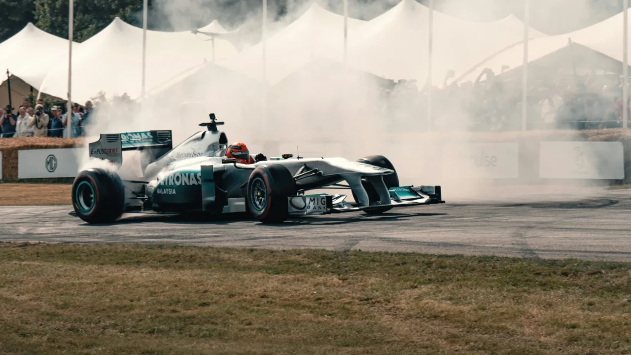 A silver Formula 1 race car performs a smoky drift on a paved track with tire smoke billowing around it, and spectators watching from behind the barriers under white canopy tents in the background.
