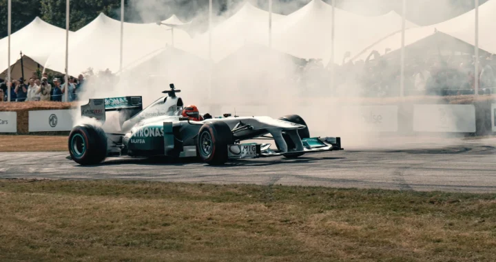 A silver Formula 1 race car performs a smoky drift on a paved track with tire smoke billowing around it, and spectators watching from behind the barriers under white canopy tents in the background.