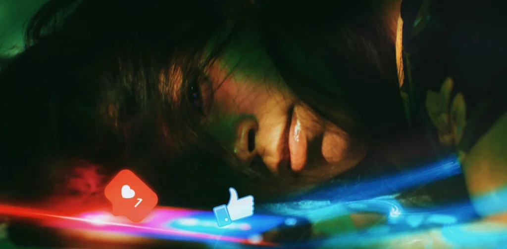 A young woman lying on her side, looking at a smartphone, with a glowing social media like and heart icon reflected on her face.