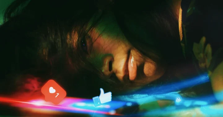 A young woman lying on her side, looking at a smartphone, with a glowing social media like and heart icon reflected on her face.