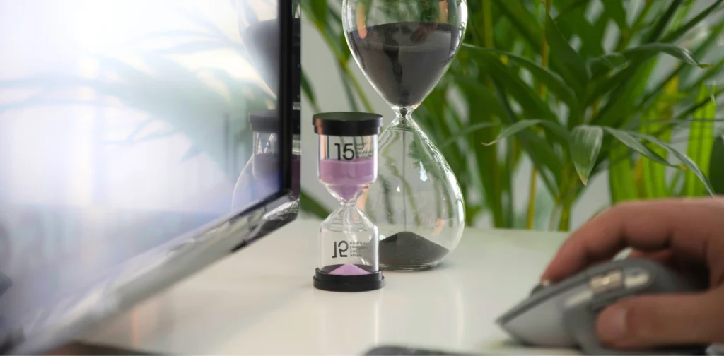 two hourglasses on a desk with a desktop screen and a hand on a mouse