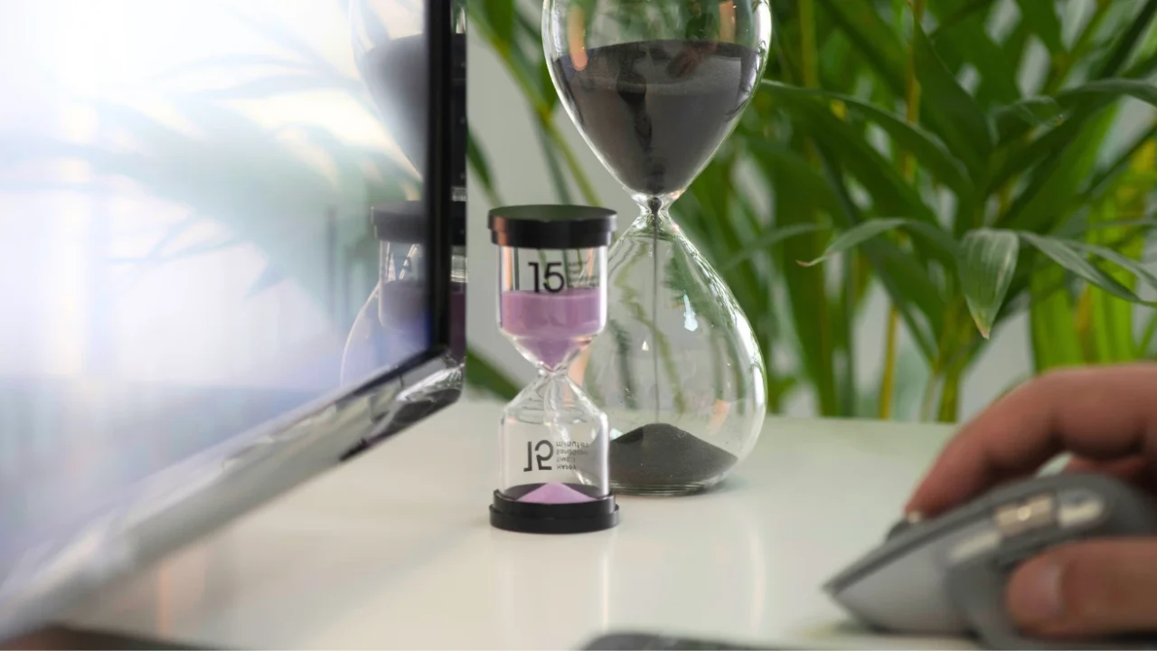 two hourglasses on a desk with a desktop screen and a hand on a mouse