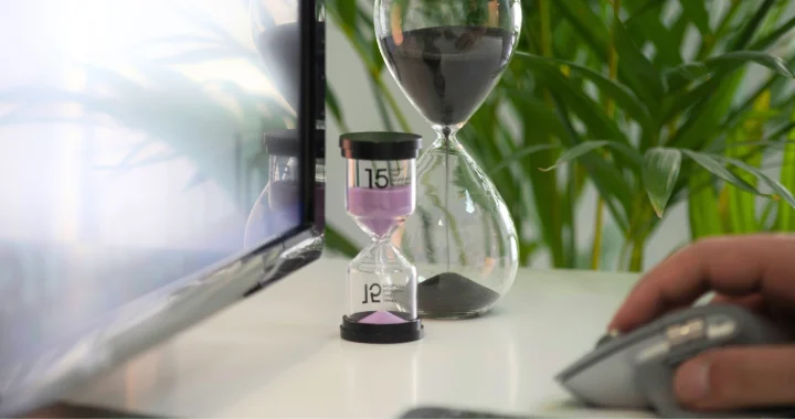 two hourglasses on a desk with a desktop screen and a hand on a mouse