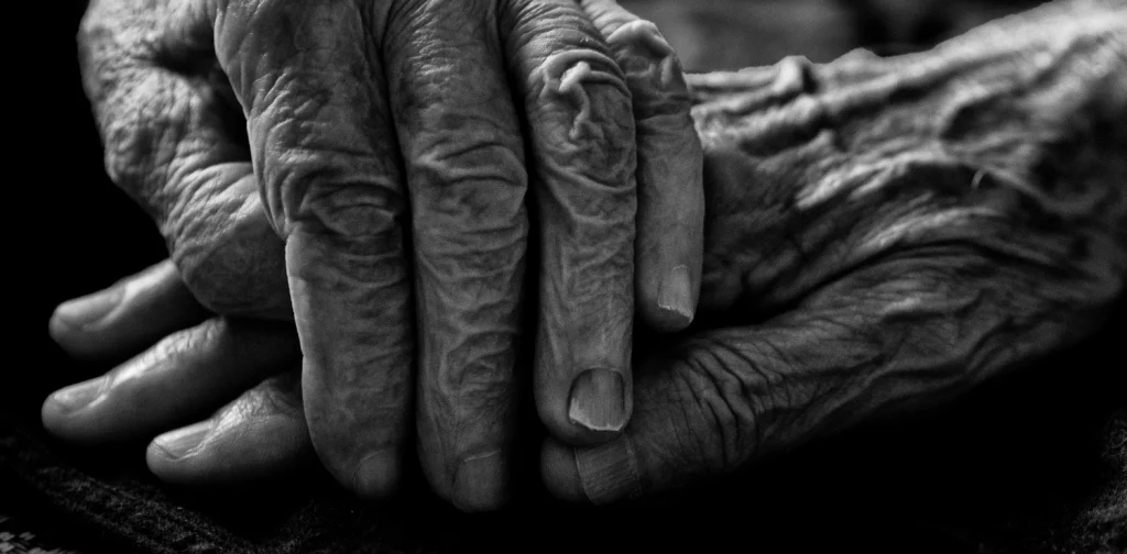Close-up black-and-white image of an elderly person’s wrinkled hands resting together in their lap.